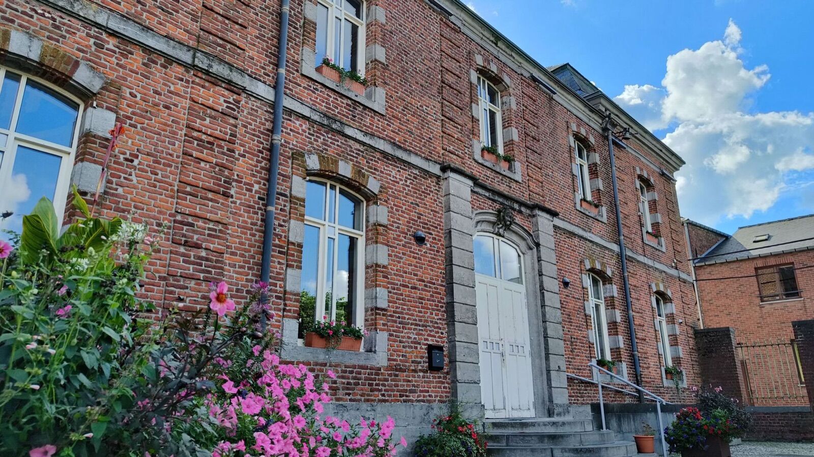Bâtiment communal de Silly en briques rouges aux encadrements de pierre, fleuri de géraniums roses sous un ciel bleu