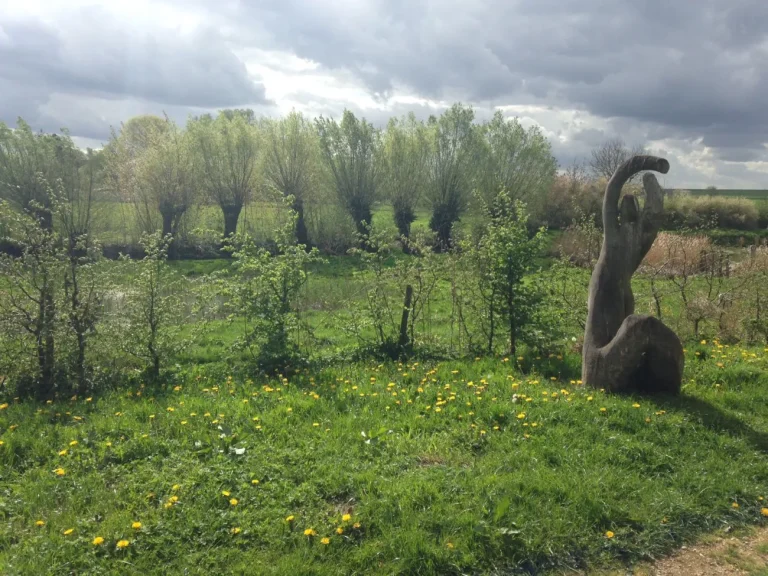 Sculpture en bois dans une prairie fleurie, devant une rangée de saules têtards