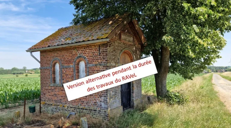 Petit bâtiment en briques au toit de tuiles sous un tilleul, au bord d'un chemin de campagne, avec la mention « Version alternative pendant la durée des travaux du RAVeL »