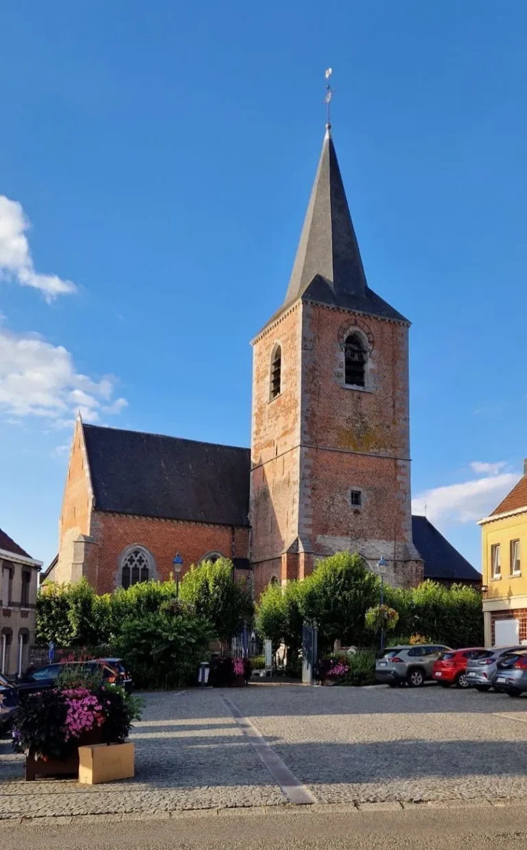 Église de briques au haut clocher d'ardoise, sur une place pavée fleurie