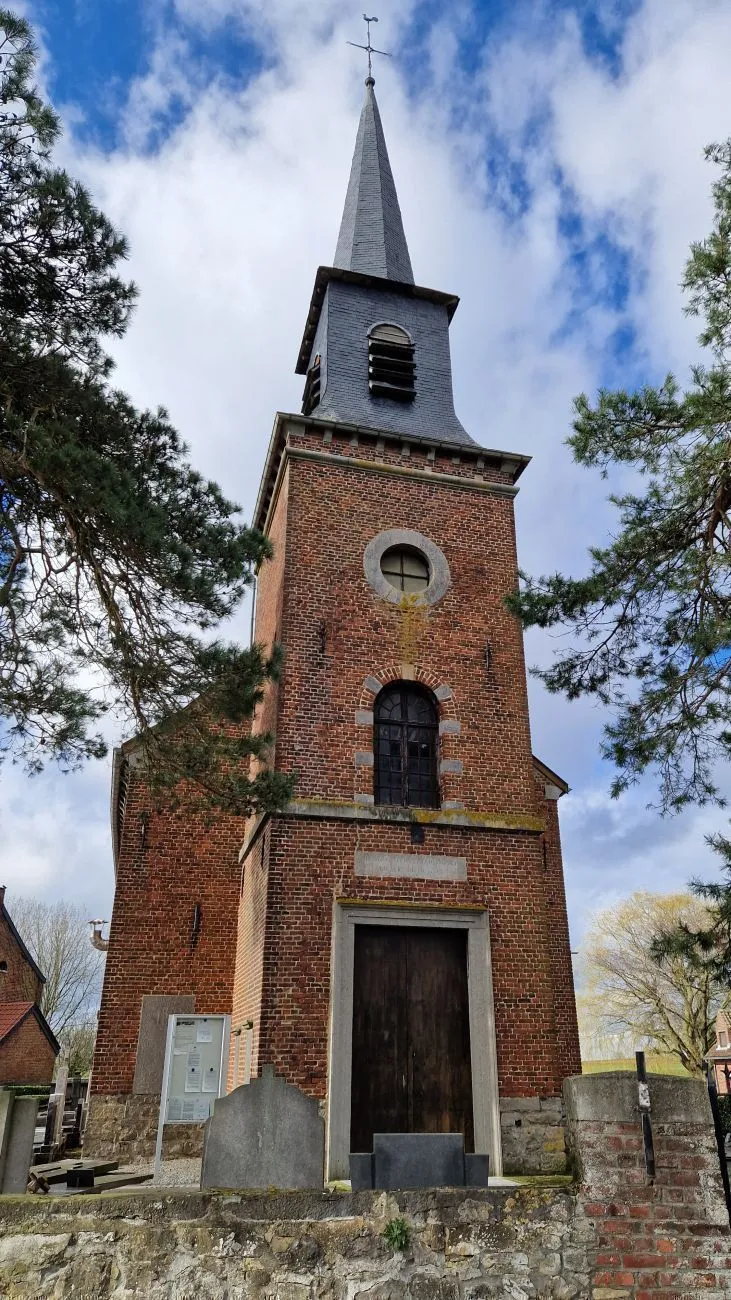 Tour-clocher en briques d'une église au portail en bois, entre deux conifères