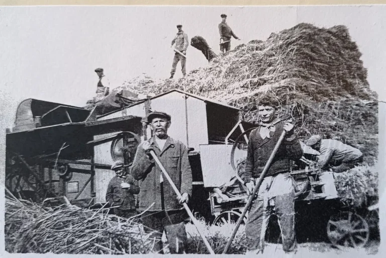 Photo ancienne en noir et blanc : des ouvriers agricoles posent devant une batteuse et une meule de paille