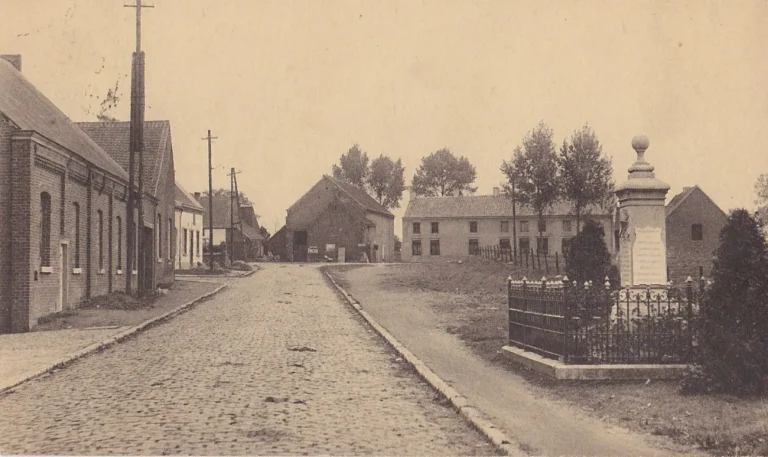 Carte postale ancienne : rue pavée bordée de maisons, avec un monument entouré d'une grille