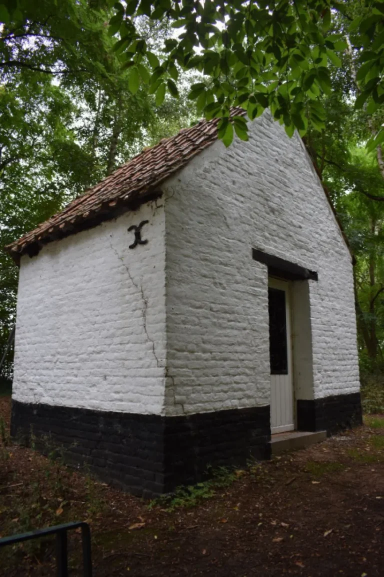 Chapelle en briques blanchies à soubassement noir, toit de tuiles et porte blanche, sous les arbres