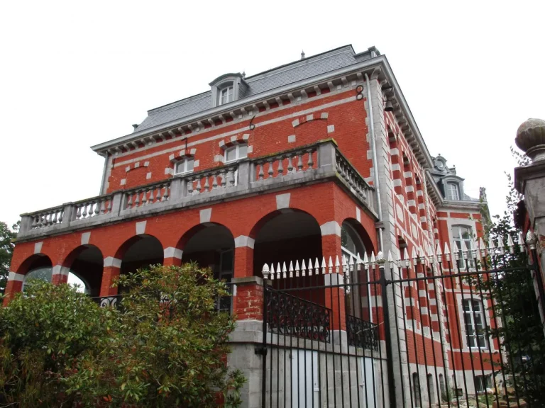 Façade de briques rouges et de pierre blanche d'une grande demeure à arcades, derrière une grille en fer forgé