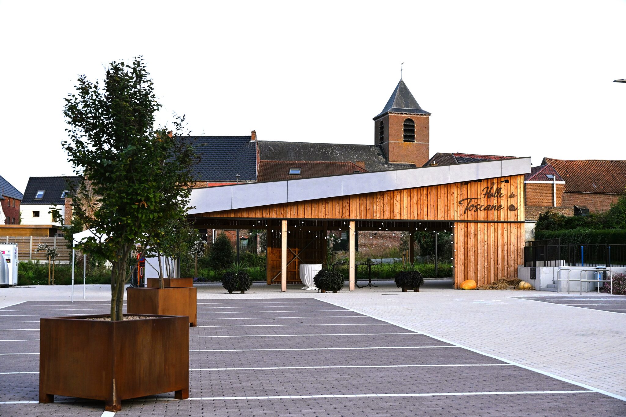 Halle de Toscane et clocher de l’église, vus depuis le parking planté d’arbres en bacs d’acier