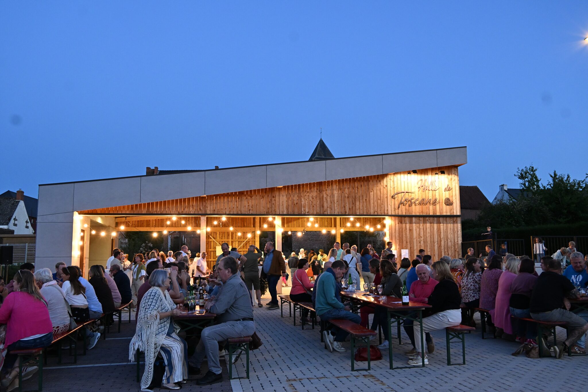 Guinguette de la Halle de Toscane au crépuscule : habitants attablés sur des bancs devant la halle en bois illuminée de guirlandes